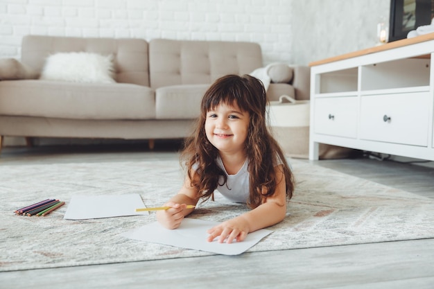 Ein süßes kleines Mädchen liegt auf dem Boden des Hauses und zeichnet mit Bleistiften die Freizeit der Kinder