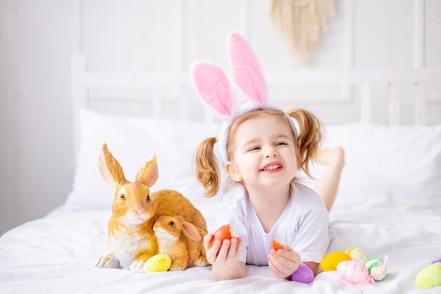 Ein süßes kleines Mädchen mit Hasenohren und bunten Eiern auf einem weißen Bett zu Hause spielt und lächelt ein blondes Kind feiert fröhliche Ostern