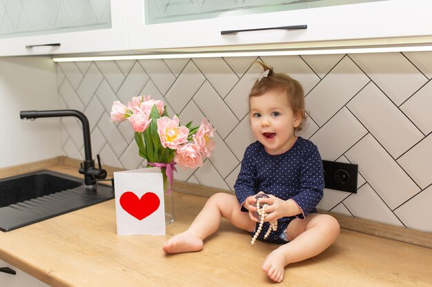 Ein süßes kleines Mädchen sitzt in der Küche mit einem Strauß rosa Tulpen, einer schönen Geschenkpostkarte