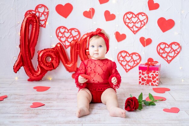 Ein süßes kleines Mädchen sitzt in einem roten Bodysuit vor einem Hintergrund aus roten Herzen und die Inschrift liebt das Konzept des Valentinstags