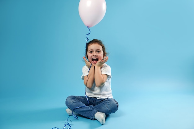 Ein süßes kleines Mädchen sitzt mit einem Ballon in den Händen auf einer blauen Oberfläche und freut sich über die Überraschung ihres Geburtstages. Ausdruck freudiger Gefühle