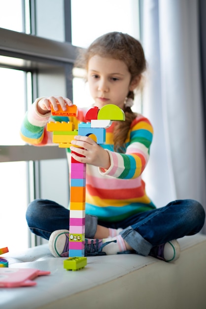 Ein süßes kleines Mädchen spielt Baukasten. Kindheit. Entwicklung. Sie sitzt in der Nähe eines Fensters mit einem großen Teddybären. Das Mädchen trägt einen farbigen Pullover und Jeans.