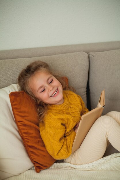 Ein süßes kleines Mädchen von vier Jahren in einem gestrickten orangefarbenen Pullover liest ein Buch in einem gemütlichen Zimmer.