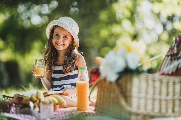Ein süßes lächelndes kleines Mädchen, das einen Frühlingstag in der Natur verbringt und Orangensaft beim Picknick trinkt.