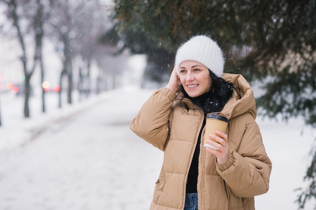 Ein süßes Mädchen hält eine Einwegtasse Kaffee und passt im Winter ihren Hut an