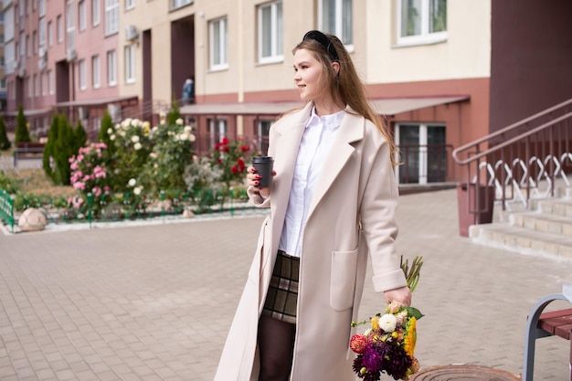 Ein süßes Mädchen in einem Mantel hält ein Glas Kaffee und einen Blumenstrauß auf der Straße
