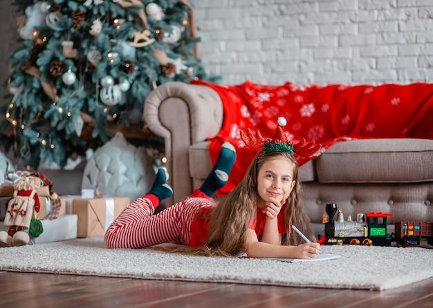 Ein süßes Mädchen in einer Weihnachtsmütze schreibt einen Brief an den Weihnachtsmann in der Nähe des Weihnachtsbaums Glückliche Kindheit