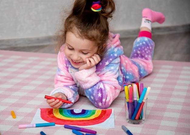 Ein süßes Mädchen in Regenbogenkleidung zeichnet einen Regenbogen mit bunten Markern