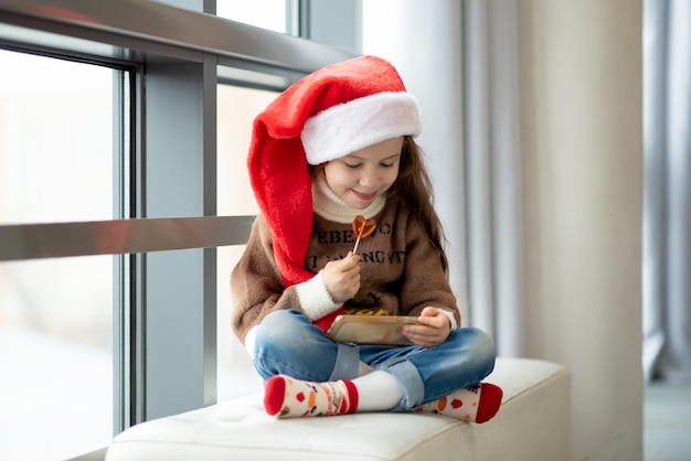 Ein süßes Mädchen in Santa's Hut hält eine Süßigkeit in Form eines Herzens in ihren Händen Neujahr Weihnachten