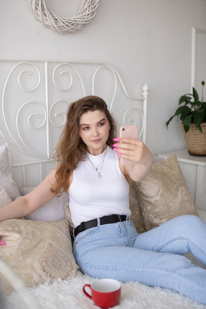 Ein süßes Mädchen sitzt mit einem Telefon auf einem weißen Bett und macht ein Selfie
