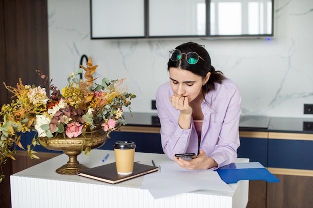 Ein süßes Mädchen steht neben einem Tisch mit Blumen und schaut auf ihr Telefon. Von zu Hause aus arbeiten