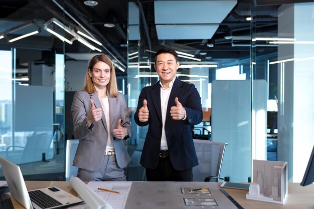 Ein Team erfolgreicher Architekten, ein asiatischer Mann und eine Frau, freuen sich über den erfolgreichen Abschluss des Projekts, schauen in die Kamera und halten den Daumen hoch, lächeln glücklich und freuen sich über die Geschäftspartner