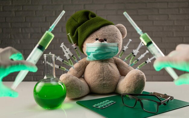 Ein Teddybär in medizinischer Maske mit Spritzen in der Schulter auf dem Arzttisch erhält eine zweite Impfspritze gegen das Corona-Virus