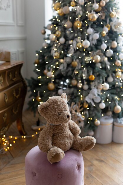 Ein Teddybär sitzt vor einem Weihnachtsbaum mit weißen Lichtern darauf.