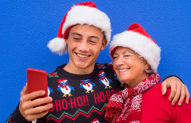 Ein Teenager-Enkel umarmt seine Großmutter und lächelt, während er vor einer blauen Wand steht und einen Weihnachtspullover und eine Weihnachtsmütze trägt. Konzept von Familie, Freundschaft und Spaß