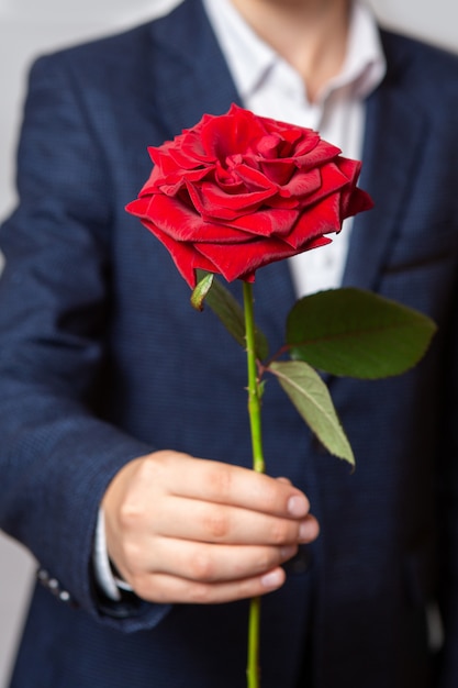 Ein Teenager hält eine rote Rose in der Hand. Der Junge trägt einen blauen Anzug und ein weißes Hemd. Nahansicht.