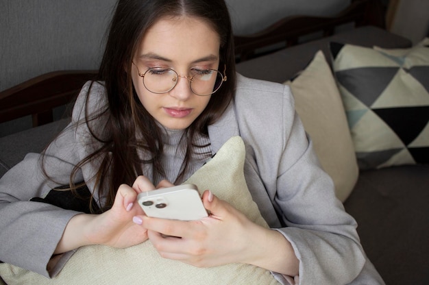 Ein Teenager sitzt mit Kissen auf der Couch und schaut auf das Smartphone