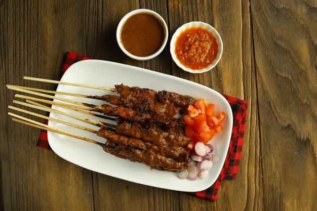 Ein Teller mit Chicken Satay, serviert auf einem weißen Teller mit geschnittenen Zwiebeln, Tomaten und einer Schüssel Sambal