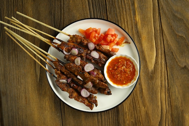 Ein Teller mit Chicken Satay, serviert auf einem weißen Teller mit geschnittenen Zwiebeln, Tomaten und einer Schüssel Sambal