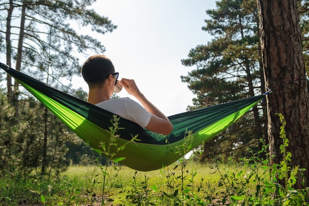 Ein Tourist in einer Hängematte ruht sich im Wald aus und trinkt aus einer Tasse.