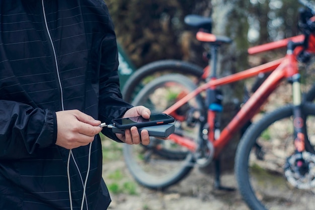 Ein Tourist lädt ein Smartphone mit einer Powerbank vor dem Hintergrund eines Fahrrads in der Natur auf