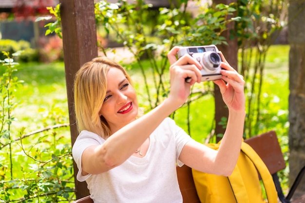 Ein Touristenmädchen, das im Sommerurlaub mit einer Kamera in einem Park fotografiert und einen Hut trägt, der auf einer Bank sitzt