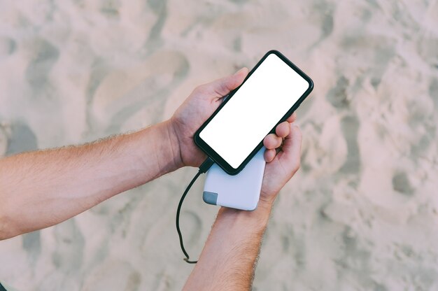 Ein tragbares Ladegerät in den Händen eines Mannes auf dem Platz des Strandsandes. Powerbank lädt das Telefon auf.