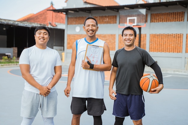 Ein Trainer und zwei lächelnde Basketballspieler mit einem Klemmbrett und einem Basketball