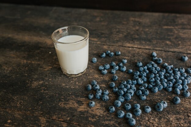 ein transparentes Glas mit Milch. Blaue frische Blaubeeren sind auf einem alten braunen Holztisch verstreut