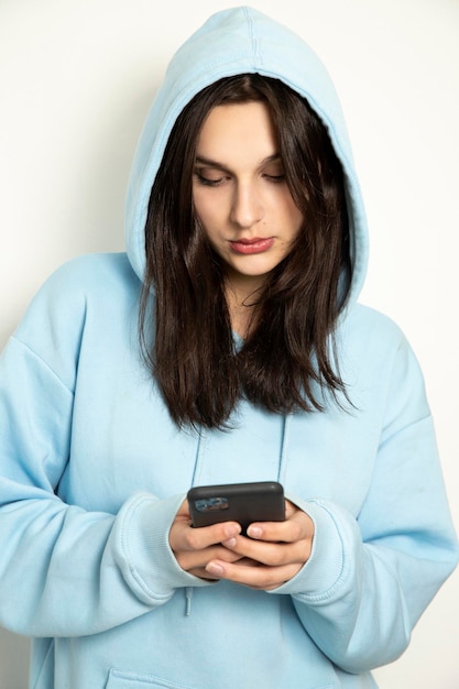 Foto ein trauriges mädchen mit einem telefon schreibt eine sms ein schönes brünettes modell in einem blauen pullover mit kapuze auf weißem hintergrund hält ein telefon in den händen