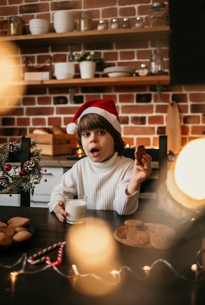 Ein überraschter kaukasischer Junge sitzt am Küchentisch in einem weißen Pullover und einer roten Weihnachtsmütze mit Milch und Keksen