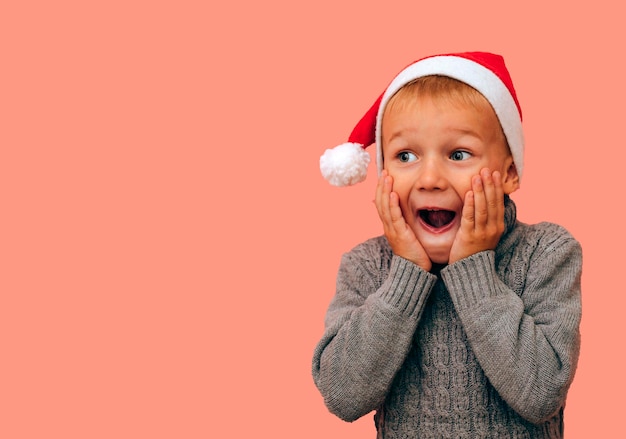 Foto ein überraschtes kind, das in der weihnachtsmütze des weihnachtsmanns wegschaut, zeigt freudige überraschung, eine wahrhaft erstaunte reaktion auf unerwartete neuigkeiten. erhobene augenbrauen und hände in der nähe des gesichts
