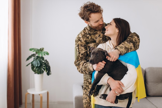 Ein ukrainisches Paar, ein Soldat in Militäruniform und ein Mädchen, das in eine ukrainische Flagge gehüllt ist, halten glücklich zusammen einen Hund in ihren Armen
