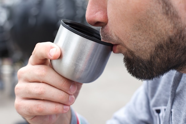 Ein unrasierter Mann trinkt Kaffee oder Tee aus einer Thermoskanne, ganz nah.