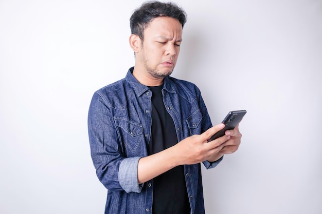 Ein unzufriedener junger asiatischer Mann sieht verärgert aus und trägt ein marineblaues Hemd mit gereizten Gesichtsausdrücken, das sein Telefon hält