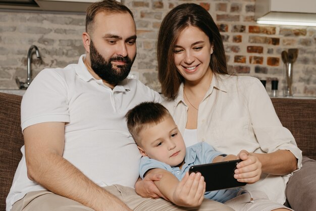 Ein Vater, ein Sohn und eine Mutter schauen sich auf dem Sofa ein Video an. Ein Kind demonstriert abends zu Hause die Show auf dem Smartphone der lächelnden Mutter und dem Vater.