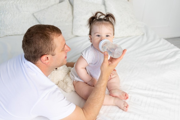 Ein Vater füttert seine kleine Tochter aus einer Flasche auf einem weißen Bett zu Hause mit dem Konzept der Babynahrung und Kinderbetreuung