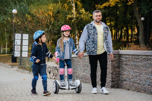 Ein Vater hilft und bringt seinen kleinen Kindern bei, während des Sonnenuntergangs Segway im Park zu fahren. Familienurlaub im Park.