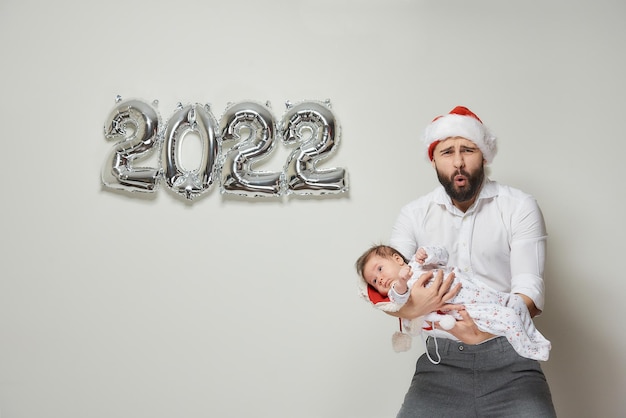 Ein Vater in einer Weihnachtsmütze aus rotem Samt hält seine kleine Tochter in der Nähe von silbernen Ballons in Form von 2022. Ein bärtiger Vater mit seinem kleinen Kind kräuselt bei einer Neujahrsparty seine Lippen zu einer Röhre.