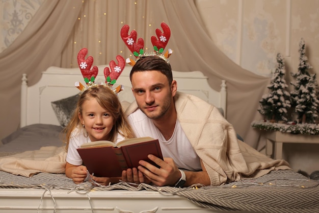 ein Vater liegt auf einem Bett mit einer kleinen Tochter in einem Hirschgeweihrand und liest ein Buch Weihnachten