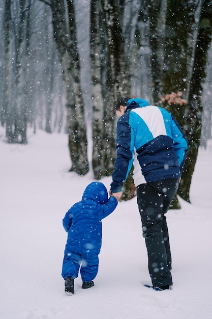 Ein Vater mit einem kleinen Kind geht unter einem Schneefall Hand in Hand durch den Schnee im Wald.