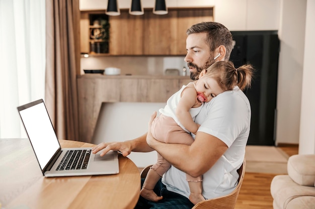 Ein Vater schläft sein Baby ein, während er am Laptop an einem Projekt arbeitet