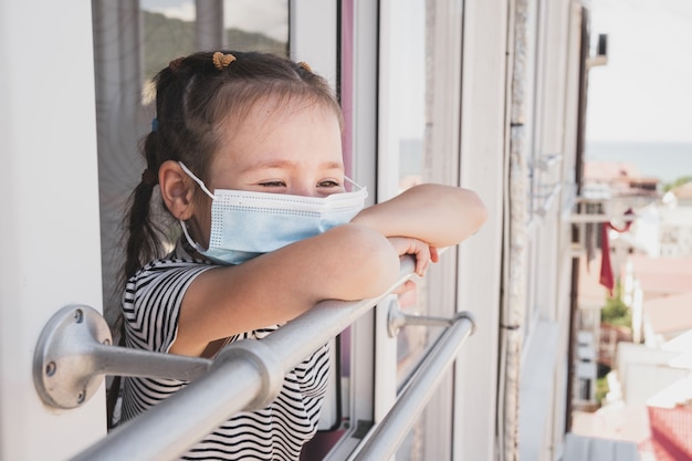 Ein verärgertes Kind steht an einem offenen Fenster das Kind ist im Urlaub erkrankt und traurig ein Mädchen asiatischer Herkunft...