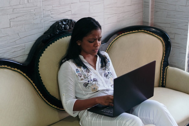 Ein verärgertes Mädchen arbeitet zu Hause, sitzt auf der Couch und schaut auf einen Laptop.