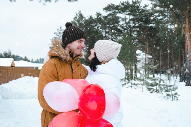 Ein verliebter Mann und eine Frau bei einem Outdoor-Date im Winter im Schnee mit einem Geschenk aus rosa und roten Luftballons in Form eines Herzens. Valentinstag, Liebe, glückliches verliebtes Paar auf einem Spaziergang