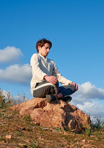 Ein vertikaler Schuss eines kaukasischen Mannes aus Spanien, der auf einem Felsen sitzt, der von Gras umgeben ist und auf Himmelhintergrund meditiert