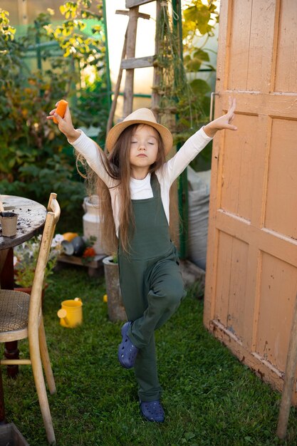 Ein vertikales Porträt eines süßen, lustigen, blauäugigen Bauernmädchens in einem Hut mit langen Haaren und einem grünen Overall Gartenbau-Hobby-Lifestyle