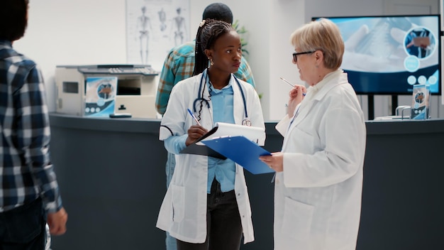 Ein vielfältiges Team von Gesundheitsspezialisten vergleicht Checkup-Dateien und Berichte in der Wartebereichslobby an der Krankenhausrezeption. Frauen mit medizinischem Fachwissen sprechen im Wartezimmer über Krankheiten.