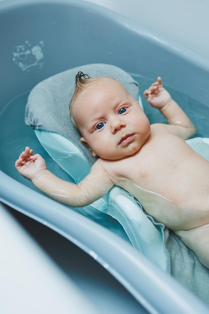 Ein viermonatiger kleiner Junge badet in einer Badewanne, während er auf einem Stand liegt und sich um ein Neugeborenes kümmert, das das Baby badet.