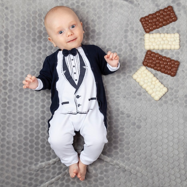 Foto ein viermonatiges baby, gekleidet in einen baby-smoking, liegt auf einem grauen teppich mit vier weißen und braunen schokoladen an seinem geburtstag, lächelnd und lachend.